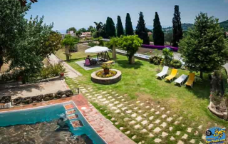 Garden view from a room at Masia Can Casadellà showing the fresh water pool, cypress and palm trees, sun loungers, with mountains and Mediterranean Sea in the background