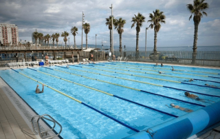 Swimmers training in separate lanes at the Olympic outdoor pool of Club Natació Atlètic-Barceloneta with the beach and sea in the background.